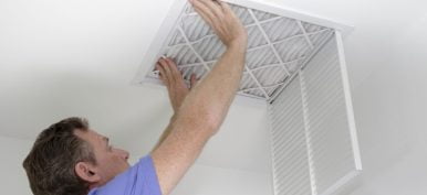 Male pushing a clean air filter into place in the ceiling with both hands. One fresh furnace air filter being secured in the intake grid of the white home ceiling.
